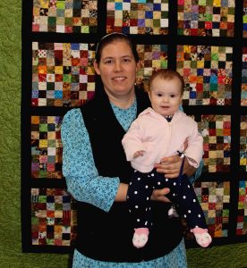 A woman holding a baby in front of a quilted wall.