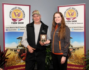 A man and woman holding a plaque in front of farm show banners.